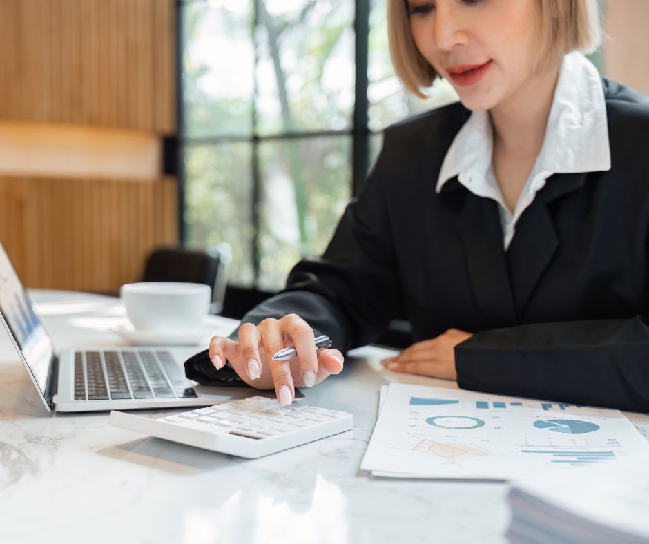 Woman sat at laptop with printouts of charts, typing into calculator